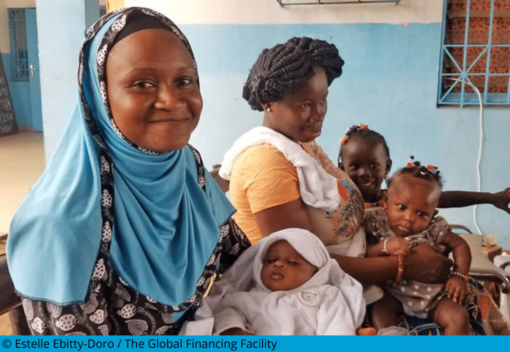 Two mothers and their three children waiting at a health center in Abobo, Côte d'Ivoire. The women who is closetst, smiles at the camera.