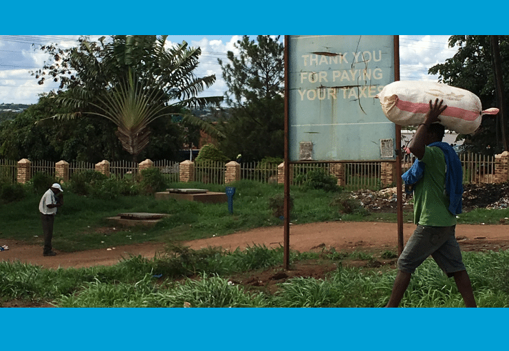 Photo of a man carrying a big bag on his head, walking by a street advertisement saying: 